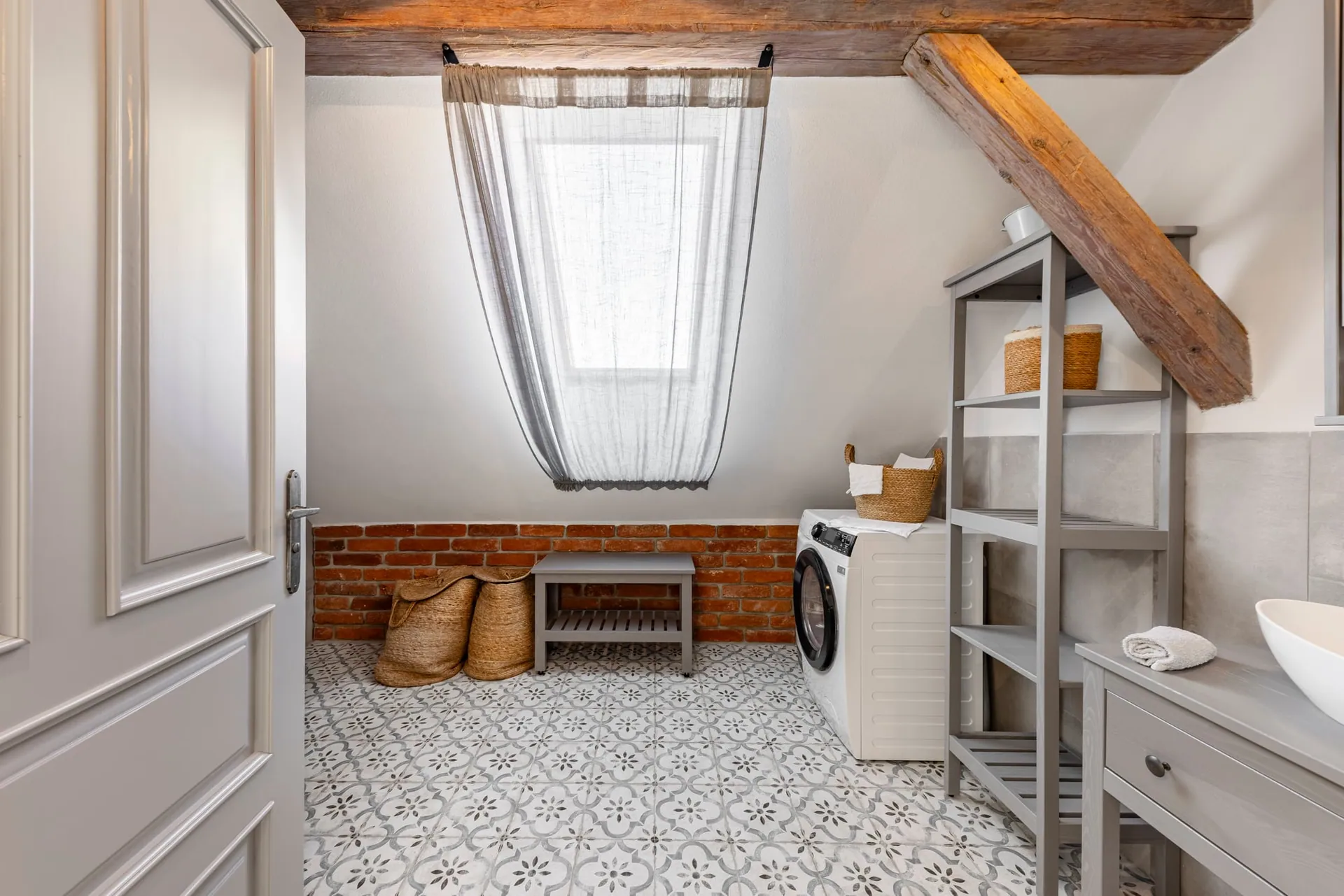 Attic bathroom with a washing machine, roof window, exposed brick band and patterned floor tiles.