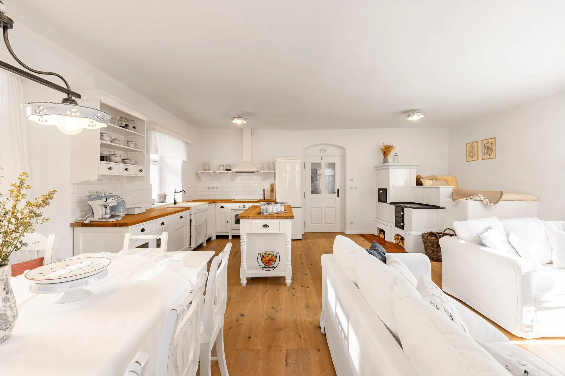 Open-plan kitchen with an island, a white tiled stove with a sleeping bench, and white sofas.