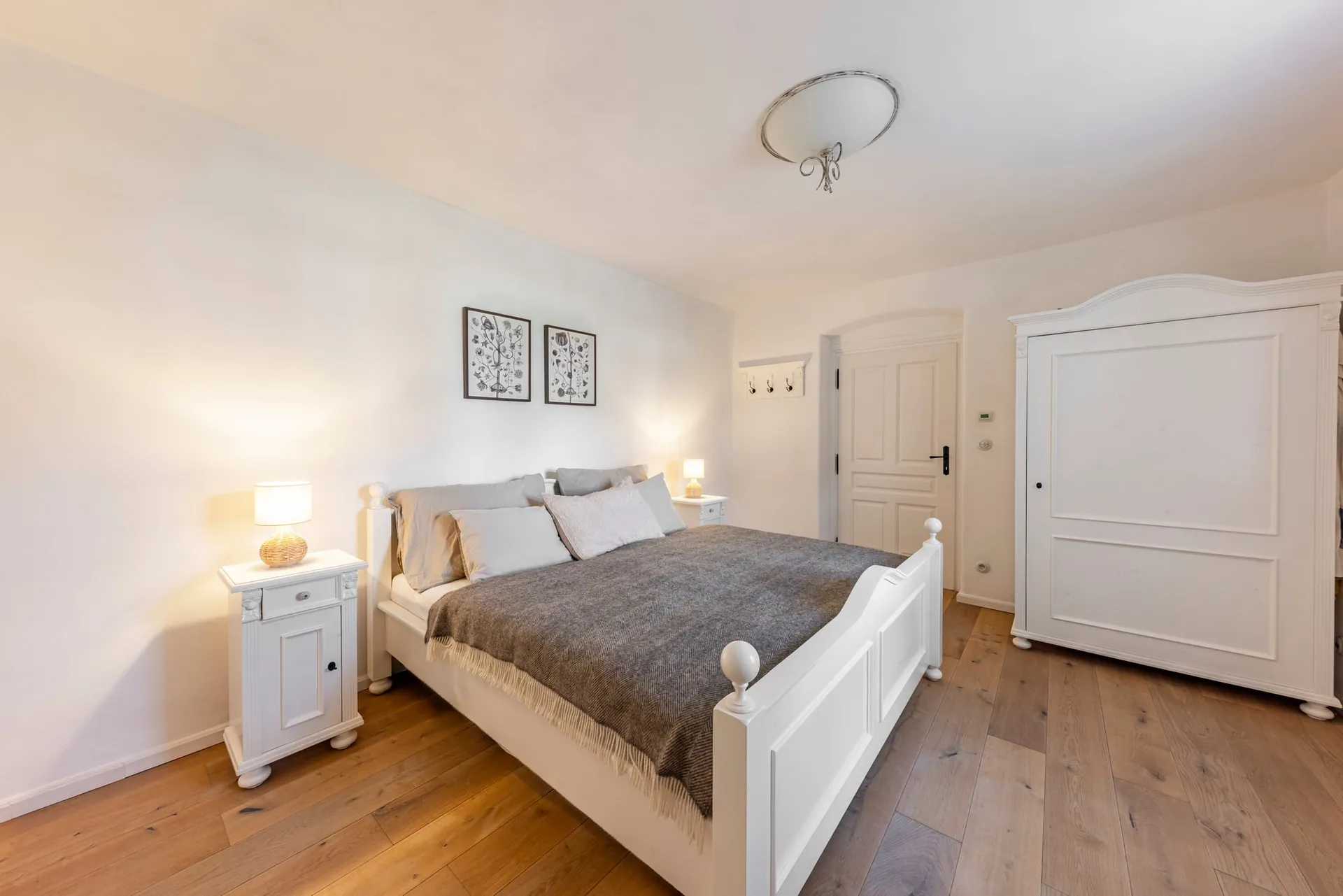 Bright bedroom with a white double bed, bedside tables with lamps and a white wardrobe.