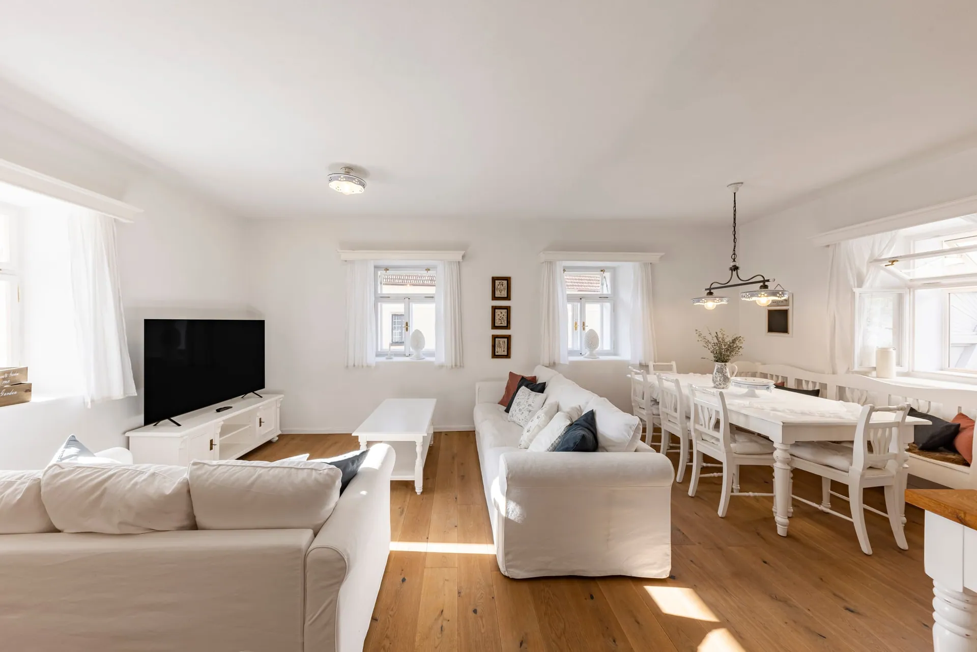Bright living room with white sofas, a TV and a dining table beside the windows.