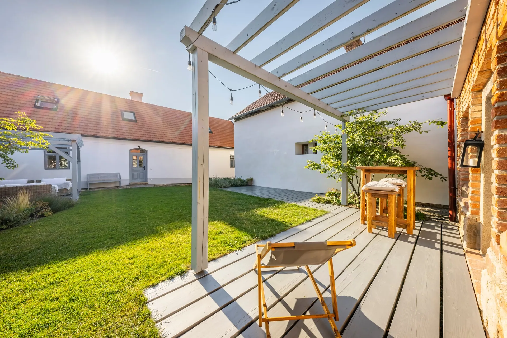 Decked terrace under the garden-house pergola, with a deckchair and the main house across the lawn.