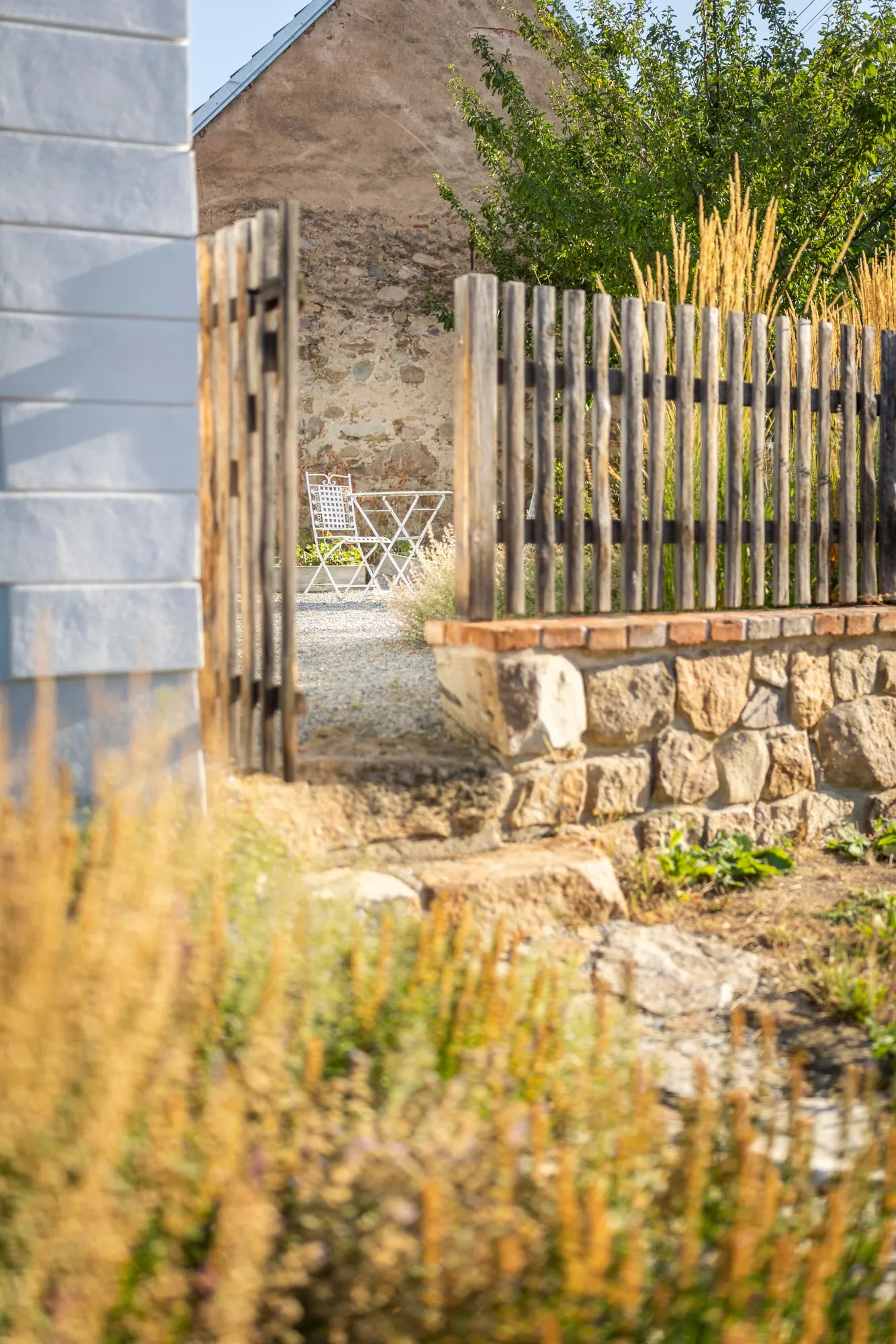 An open wooden gate in a fence on a stone base, with a white metal chair and table beyond.