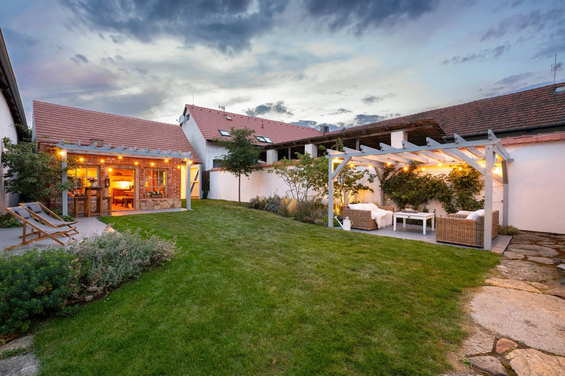 The garden at dusk: the lawn, the lit garden house and rattan seating under the pergola